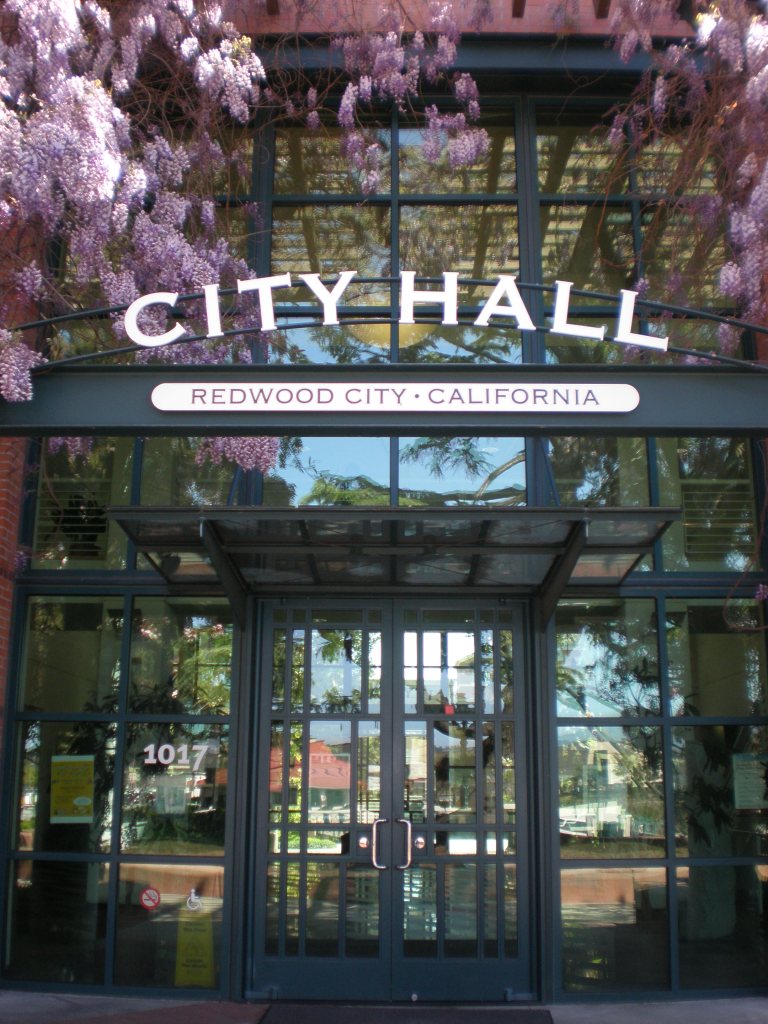 Front doors and sign, surrounded by pink cherry blossoms, of city hall in Redwood City, California.