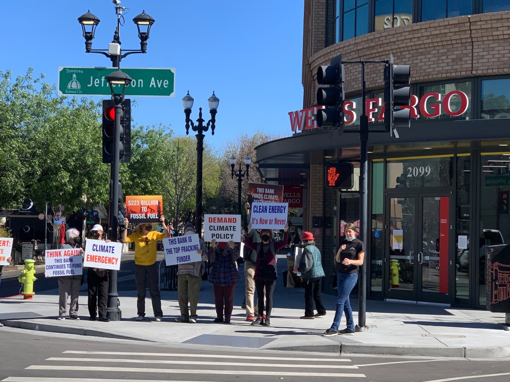 A group of climate protestors hold signs in front of a Wells Fargo bank branch location.